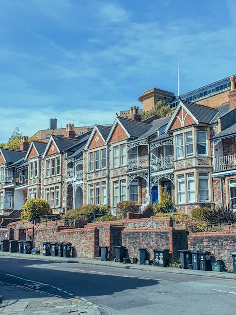 A row of Victorian-style terraced houses with ornate white ironwork balconies and decorative woodwork on the gabled roofs, set against a bright blue sky. The buildings have brick facades with light-colored window frames and small front gardens that are separated from the street by low brick walls. In the foreground, there is a paved street with several black wheelie bins lined up along the pavement, suggesting waste disposal or collection activity by a private waste management service. The scene appears to be in an urban residential area, with the houses closely packed together and some greenery visible in the form of small bushes or shrubs in front gardens. The overall lighting indicates a clear, sunny day, highlighting the architectural details and vibrant urban environment, consistent with the context of independent rubbish removal or house clearance services.