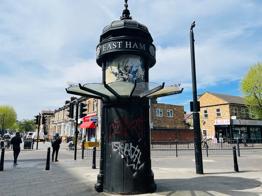 A tall, cylindrical Victorian-style street clock with a domed black metal top and decorative finial, displaying the words 'EAST HAM' on a circular sign at the top. The clock face, partially visible, is encased in glass, with a weathered appearance. Attached to the lower part of the structure is a black metal mesh panel, which has graffiti in red and white spray paint. Surrounding the clock is a paved public area with black metal bollards, some with warning signs, and a street scene in the background featuring a pedestrian crossing with four individuals walking, a red and white bus stop sign, a small shop with a red awning, residential buildings with brick facades, and a few trees with fresh green leaves. The sky is partly cloudy, with patches of blue visible, indicating daylight. The overall scene suggests an urban environment that involves regular street maintenance and cleanliness, aligning with the services of a rubbish removal company such as House Clearance Tooting.