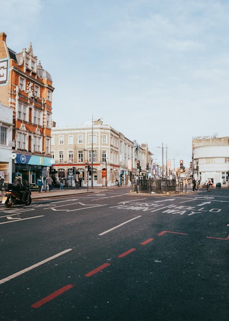 A wide-angle view of a busy Tooting street scene with a clear, bright sky overhead. On the left, there is a row of multi-storey Victorian-style buildings with ornate brickwork and decorative elements, housing various shops at street level. Pedestrians are seen walking along the pavement, some entering or exiting the shops. In the foreground, a section of road is visible with red dashed lines marking a designated lane or parking area, and white directional arrows painted on the asphalt. Across the street, there are additional buildings, including a white curved structure on the right side and other commercial premises further down the street. Several vehicles, including a motorbike and cars, are parked or moving along the road. Streetlights and traffic signals are present, and the overall atmosphere is that of a typical urban shopping and pedestrian area, with a focus on the infrastructure that supports both vehicle traffic and foot traffic. This scene exemplifies an active city centre environment suitable for private waste collection services and on-site rubbish removal efforts by companies like House Clearance Tooting.